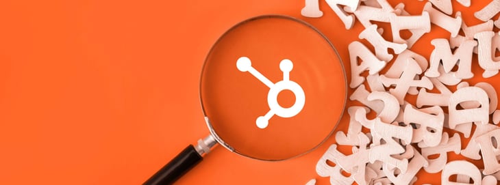 Magnifying glass highlighting the HubSpot logo on an orange background with scattered wooden letters, symbolizing HubSpot marketing terms.